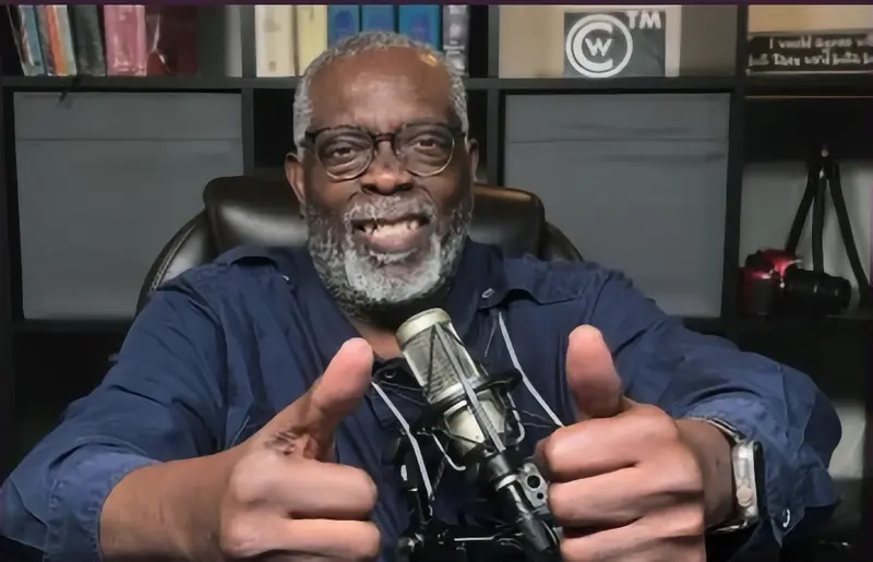 A Tony Mobley with glasses and a gray beard sits in front of a microphone, smiling and giving two thumbs up, with bookshelves in the background.